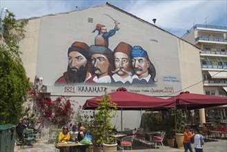 Large mural of historical figures above an urban café with passers-by, graffiti with 5 heroes of