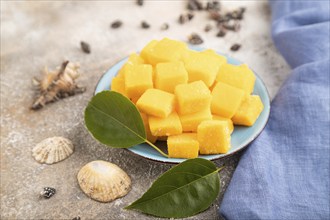 Dried and candied mango cubes on blue plate on brown concrete background and linen textile. Side