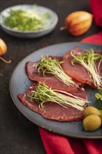 Slices of smoked salted meat with cilantro microgreen on black concrete background and red textile.