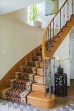 Wooden spiraling staircase with burgundy and cream coloured rug runner and brass rods leading to