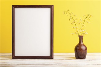 Wooden frame with yellow barrenwort flowers in ceramic vase on yellow pastel background. side view,