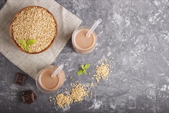 Organic non dairy oat chocolate milk in glass and wooden plate with oat seeds on a gray concrete