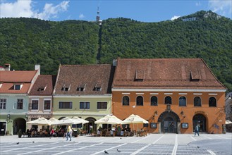 A lively town square in front of historic buildings with cafés at the foot of a green mountain,