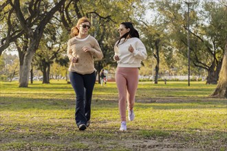 Two women jog side by side in a sunlit park, smiling as they embrace an active lifestyle.