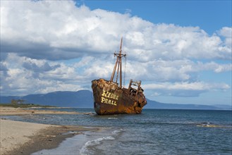 The bow of a rusty ship eats into the sandy beach against a blue sky and fluffy clouds, shipwreck