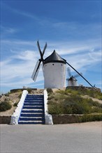 White windmill against a blue sky, flanked by a blue and white stone staircase, surrounded by