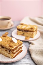 Honey cake with milk cream, caramel, almonds and a cup of coffee on a gray and pink background and