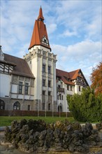 A historic building with a pointed tower and red roof, surrounded by autumn trees under a clear