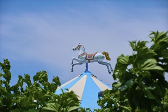 A carousel horse stands on a blue and white striped roof, surrounded by green leaves under a clear