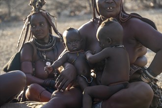 Married Himba woman with their babies in their arms sitting in front of the hut of the first woman,