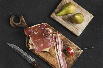 Cured duck meat on a wooden board next to two ripe pears. A knife rests nearby, highlighting the