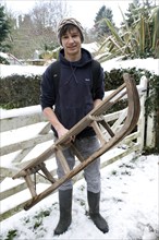 Model released portrait of teenage boy in the snow holding a wooden sledge, Suffolk, England,