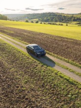 A car drives on a dirt track between large fields under a sky with clouds and evening sun, car