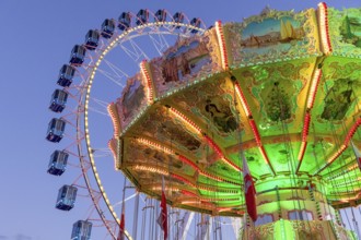 A brightly lit carousel in front of a large Ferris wheel at dusk at a funfair, Wellenflug,