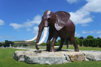 Metal sculpture of an elephant on a rock foundation in a green park under a blue sky with white