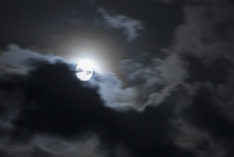 The full moon shines brightly in a cloudy, stormy, black sky and has a halo (corona),