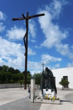 Large cross and statue of Pope John Paul II on a square with flowers and blue sky, Santuario de