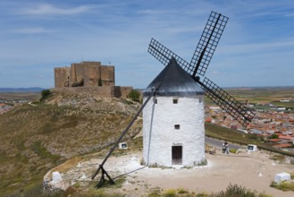 An old windmill in front of a medieval castle on a hill with a village in the background under a