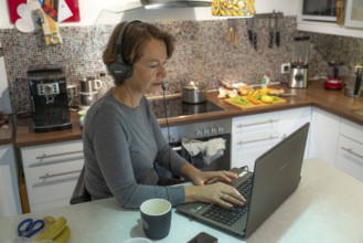 Woman, mid-50s, works from home, with laptop and communicates with colleagues via headset, home