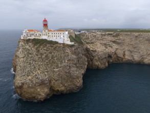 A red lighthouse and white buildings stand on an imposing cliff with a wide view of the ocean,