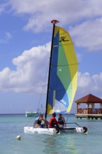 People on a colourful sailing boat in the clear sea near a jetty with a pavilion under a blue sky,