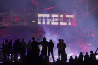 A silhouette of several festival visitors in front of the logo projected onto an excavator at the