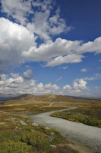 A hiking trail leads through a wide, hilly landscape towards the mountains under a blue sky,