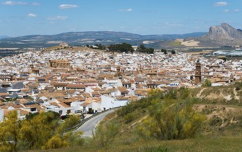 Spanish town with white buildings on a hill against a mountain backdrop under a blue sky,