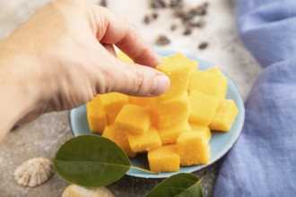 Dried and candied mango cubes on blue plate with hand on brown concrete background and linen