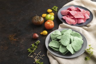 Green and red potato chips with herbs and tomatoes on black concrete background and linen textile.