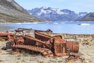 Rusty cars and barrels on a fjord in front of steep mountains, remains of a US airbase from the