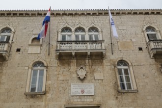 Old beige and tan stone cladded Trogir Town Hall building facade with balcony, flags, arched