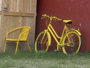 A bright yellow bicycle and matching chair placed outdoors by a wooden door and red wall,