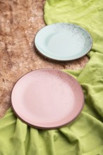 Two empty blue and pink plates on brown concrete background and green textile. Side view, close up