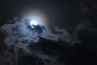 The full moon shines brightly in a cloudy, stormy, black sky and has a halo in rainbow colours