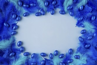 Frame made of blue and turquoise feathers with blue chocolate eggs on a light blue background