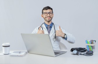 Smiling doctor sitting with laptop giving thumbs up. Male doctor with laptop showing thumbs up in