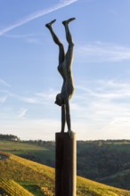 A statue of a person doing a handstand on a metal post in front of a landscape background at