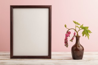 Wooden frame with pink dicentra, broken heart in ceramic vase on pink pastel background. side view,