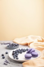 Purple macarons or macaroons cakes with blueberries on white ceramic plate on a gray and orange