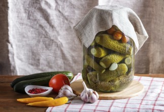 Pickled cucumbers and tomatoes in a glass jar on linen tablecloth and wooden table. Homemade, copy