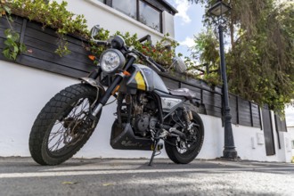 A stylish motorbike is parked on a street next to a house surrounded by green plants, Canary