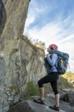 Female climber standing in front of a rock, carrying climbing rope, looking up, observing rocky