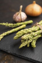 Bunch of fresh green asparagus, garlic, onion on slate board on black concrete background. Side