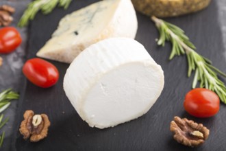 White goat cheese and various types of cheese with rosemary and tomatoes on black slate board on a