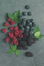 A mix of ripe berries, with leaves scattered on the table, raspberries, blackberries, blueberries,