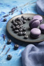 Purple macarons or macaroons cakes with blueberries on ceramic plate on a blue concrete background