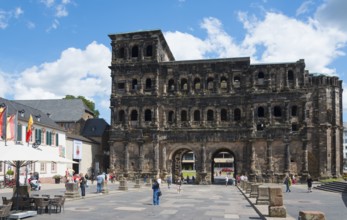 A large, ancient building with several arches and windows, surrounded by people on a sunny street,