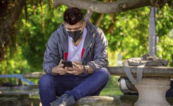 Man with a mask sitting on a bench with his phone, young man with a mask sitting with a cell phone,