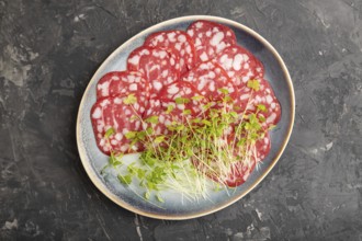 Slices of smoked cervelat salami sausage with spinach microgreen on black concrete background. Top